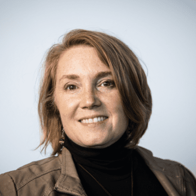 Head shot Dr Jennifer Swoyer. Jennifer is in front of a grey backdrop wearing a brown jacket with a black turtleneck