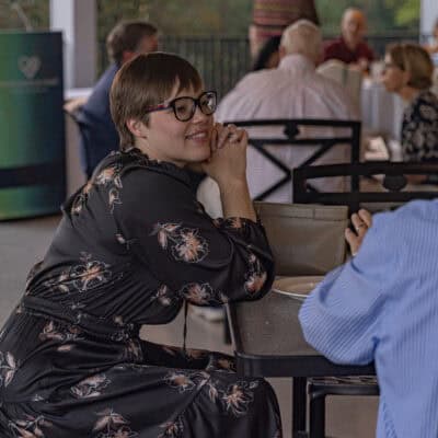 Woman wearing a floral dress seated at a table speaks to a man with grey hair