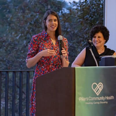 Alex Normington wearing a pattened dress stands at a podium speaking with Angela Curran standing in the background