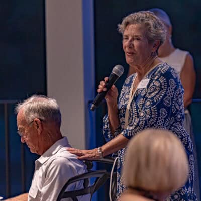 Woman stands holding a microphone with hand on the shoulder of seated man