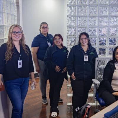 Group of 5 women stand near a desk wearing Pillars Community Health Branded clothing with a glass block wall in the background