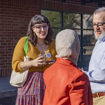 Woman wearing glasses speaks with a man in a blue button down and a woman in a red jacket wearing a grey headscarf