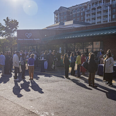 A crowd gathered outside of a brick buiding with the Pillars Community Health Logo and the sun shining