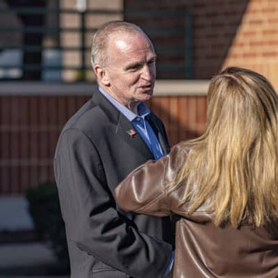 Two people stand talking to one another: A man in a suit and a person with long brown hair and a brown leather jacket facing away from the camera