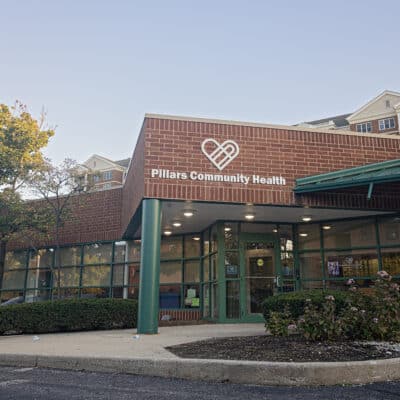 A single story brown brick and glass building with the Pillars Community Health Logo and an automatic door