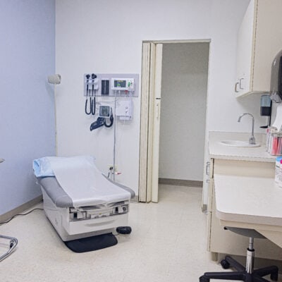 Calm medical exam room with an exam bentch medical tools on the back wall along with an entrance to another room and storage.