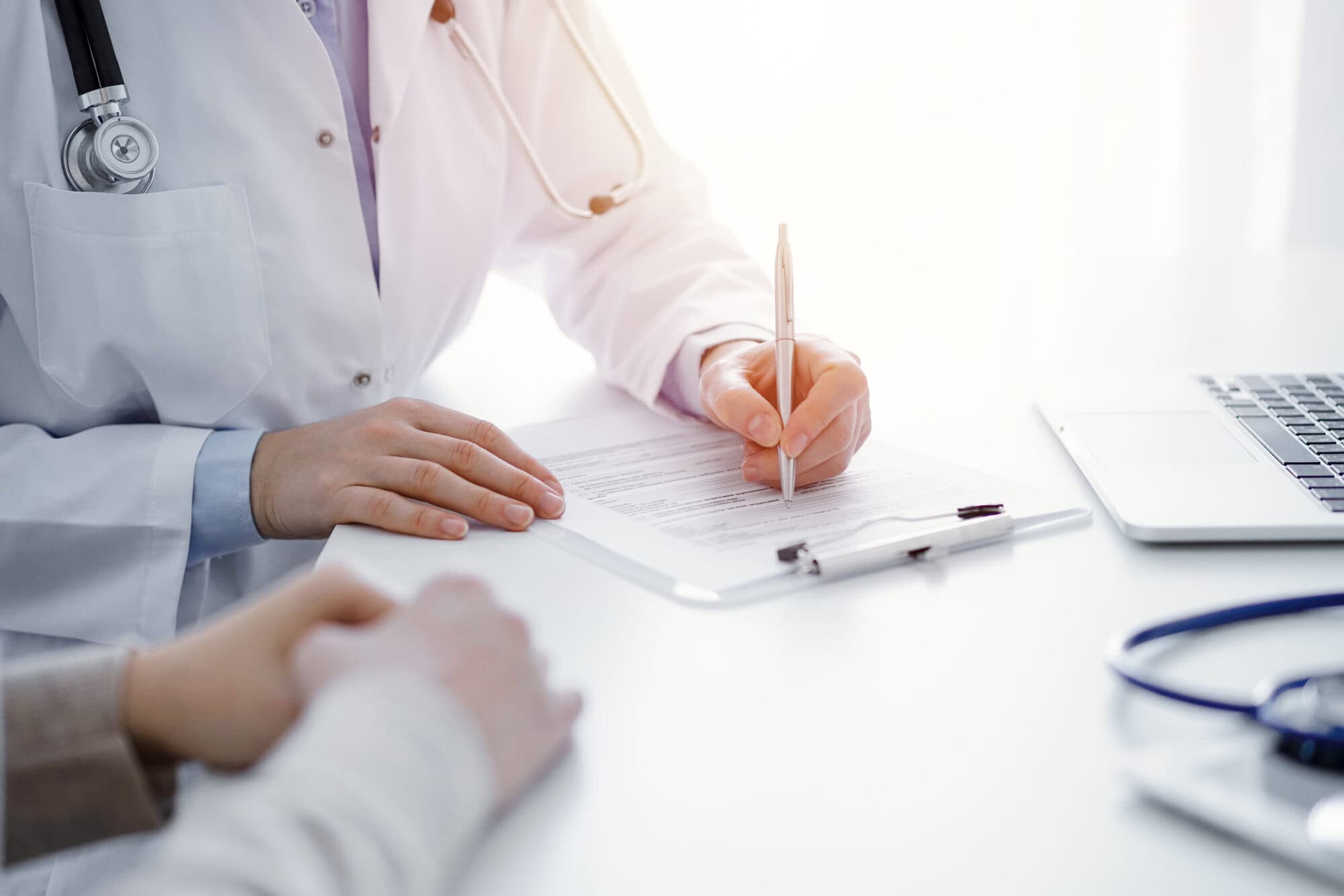 Doctor and patient sitting at the table in clinic. The focus is on female physician's hands filling up the medication history record form or checklist, close up.