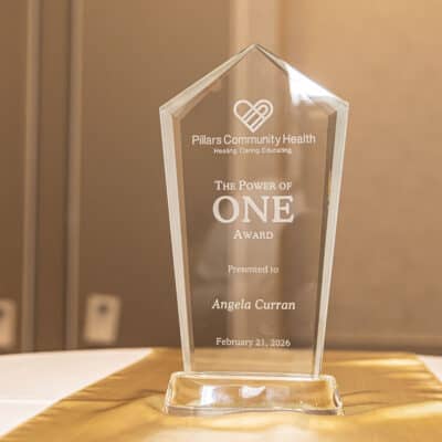 Glass award engraved with “The Power of One Award” presented to Angela Curran by Pillars Community Health on February 21, 2026.