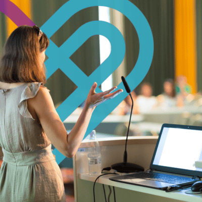 Presenter speaking to seated participants beside a laptop and microphone, with a teal and purple heart graphic overlaid.